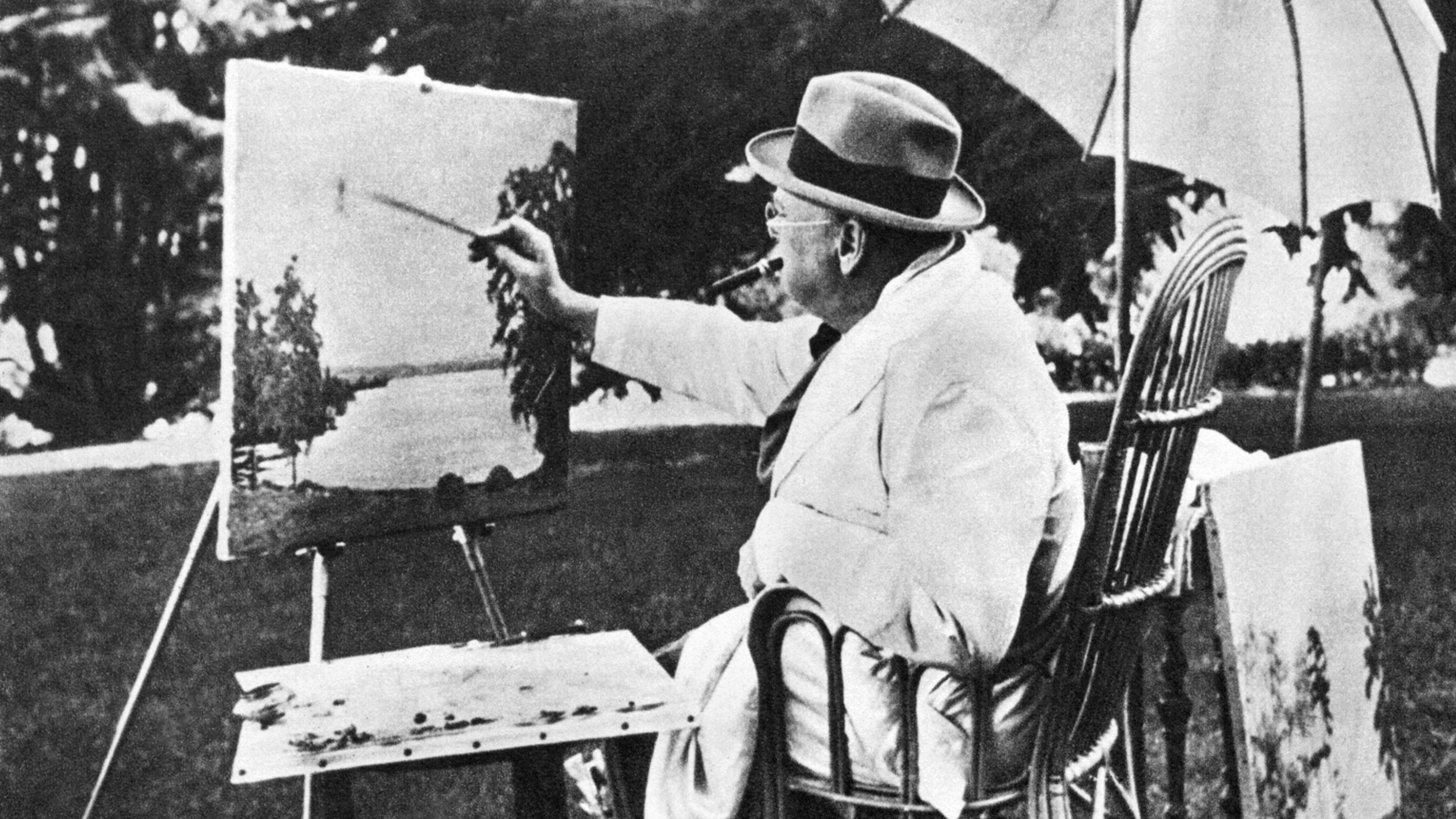 Black and white archive photo of Sir Winston Churchill seated before an easel and paint set painting beneath a parasol.