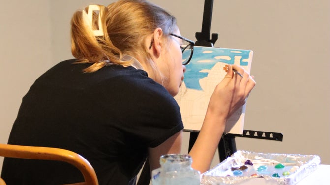 Young woman painting the sky into a landscape scene. Her canvas is propped against an easel.