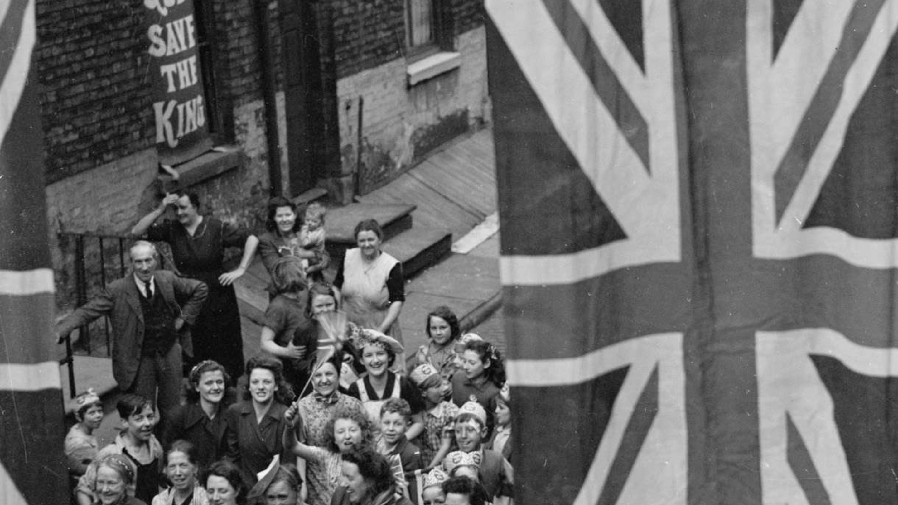 Archive photograph of VE Day celebrations at an east end street party in May1945. Men, women and children gathered in the street, smiling, holding hands, some wearing paper crowns, others waving flags beneath Union flags and bunting draped between the rows of houses on either side of the street.