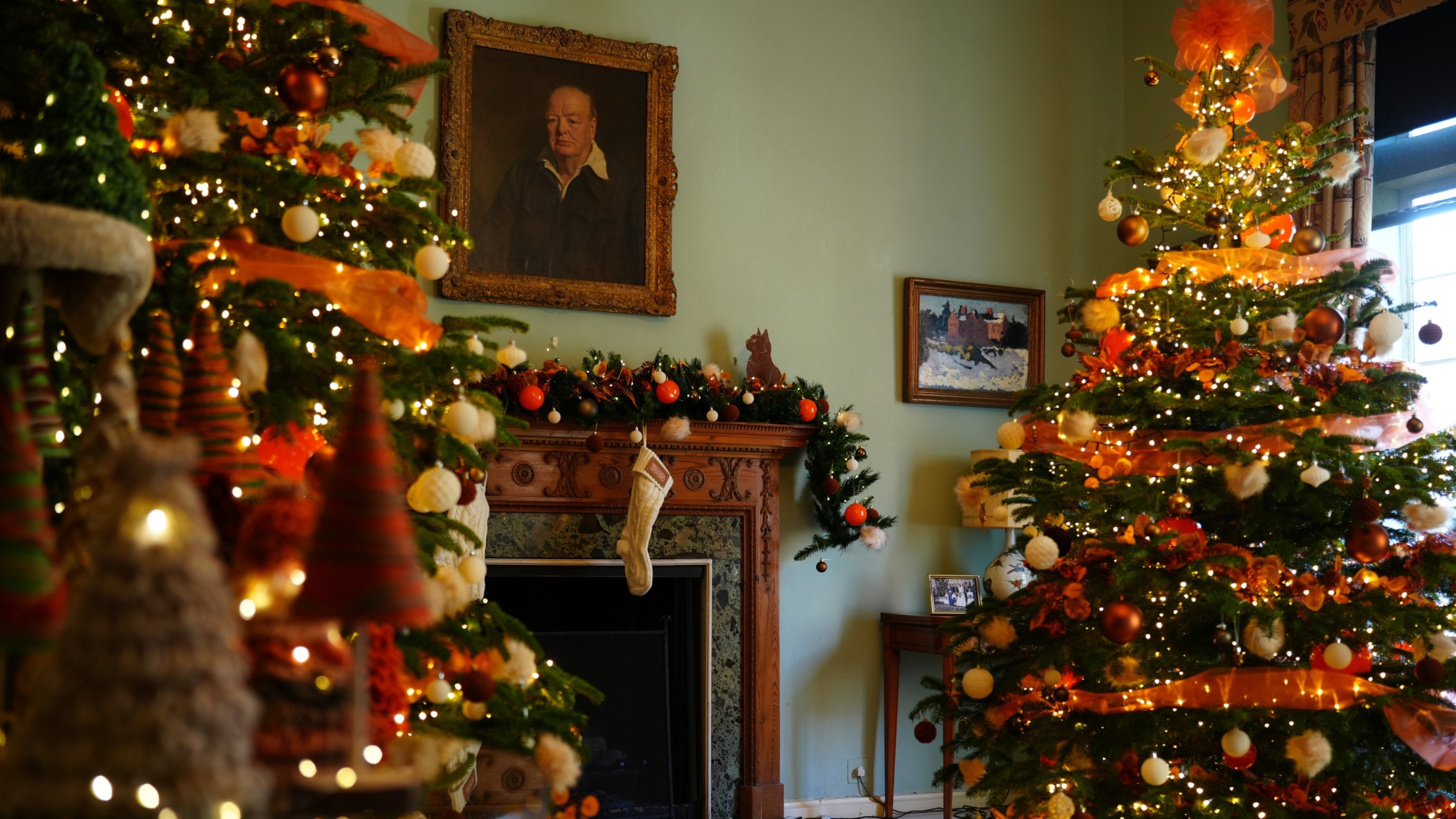 Yarn Christmas trees on a cat climbing tree, orange and white ribbons, baubles and balls of fluff decorate the Christmas Trees and garlands. A pair of white knitted stockings above the fireplace. A silhouette of a cat on the mantlepiece. Portrait of Winston Churchill on the wall and a winter landscape painting of Chartwell below.