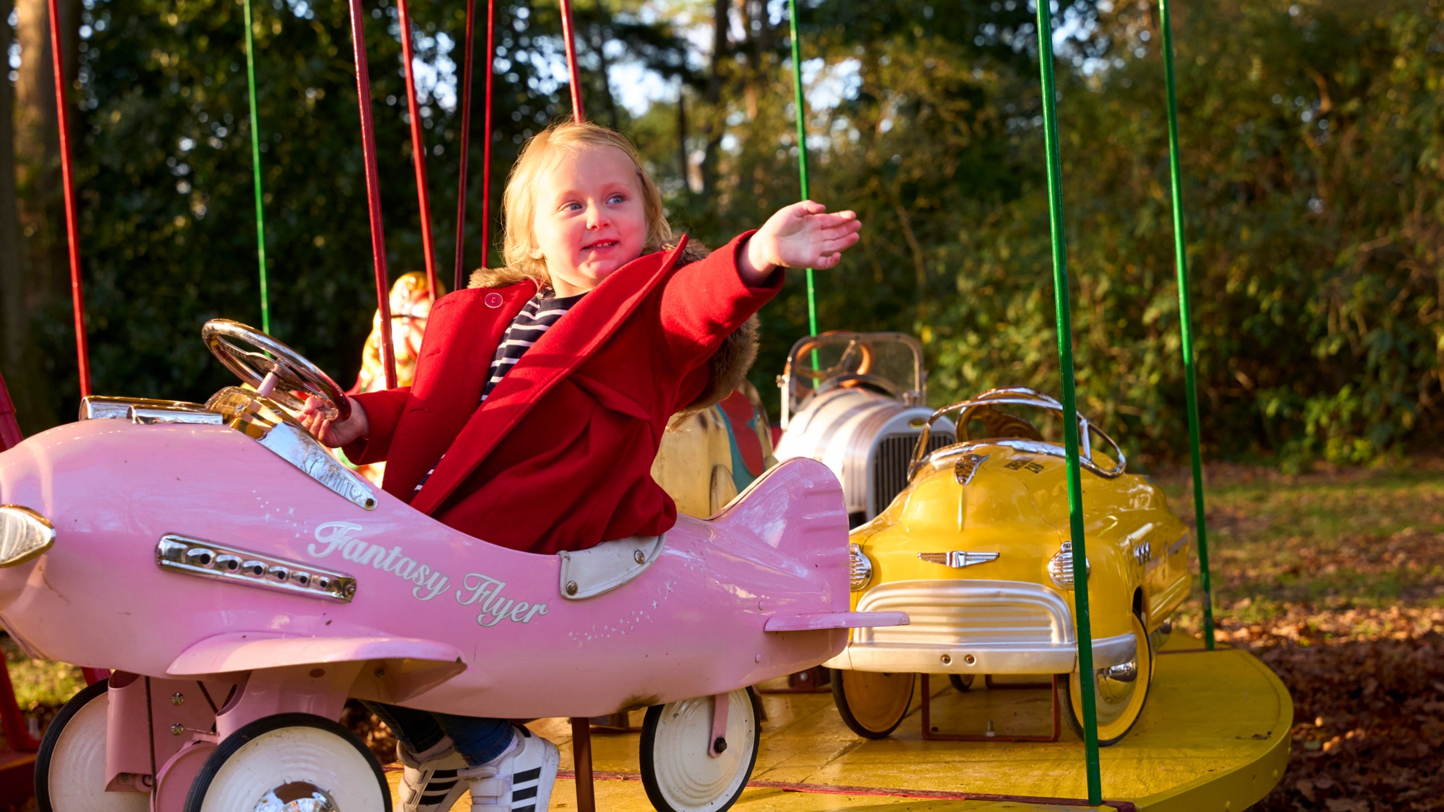 A toddler in a red coat waves whilst riding a pink airplane on a carousel.