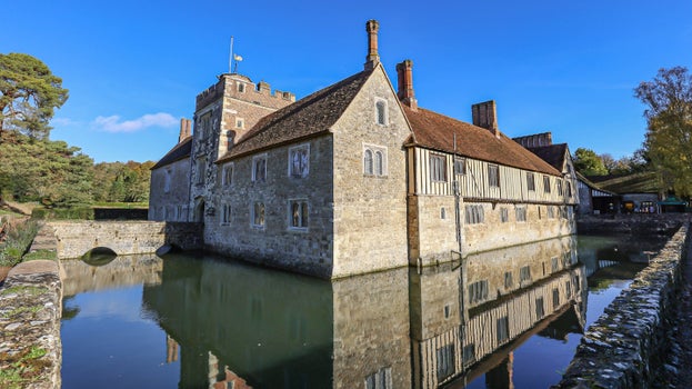 The corner of the west front of the house reflected in the moat, with a stone bridge leading to the entrance,  at Ightham Mote, Kent