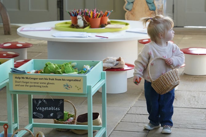 A small child holding a basket, looking away from the camera. She is standing next to a wooden green stand that has a label with the word vegetables on hanging from it.
