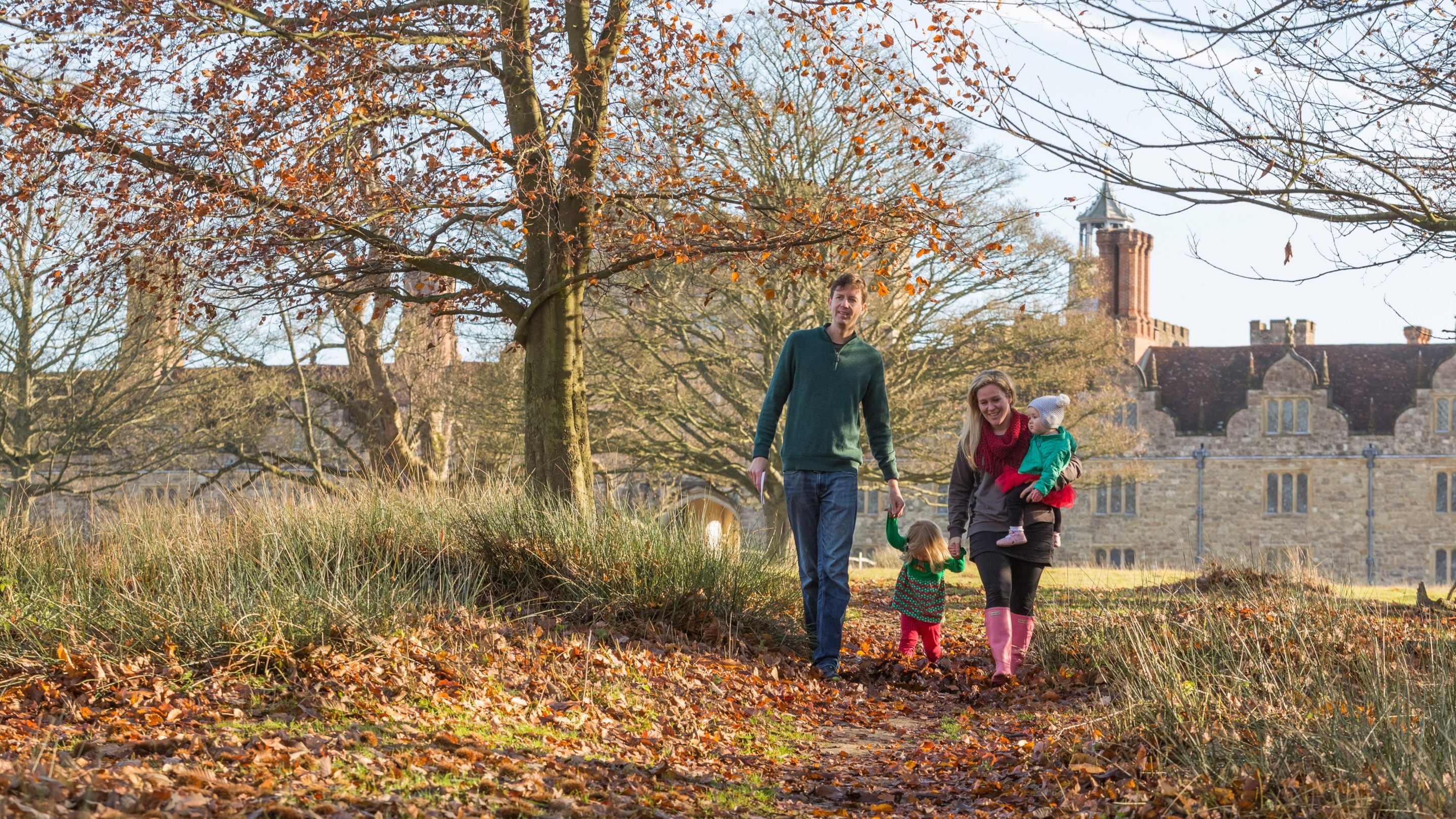 A family walks along a path covered in fallen leaves, with the house in the background at Knole, Kent