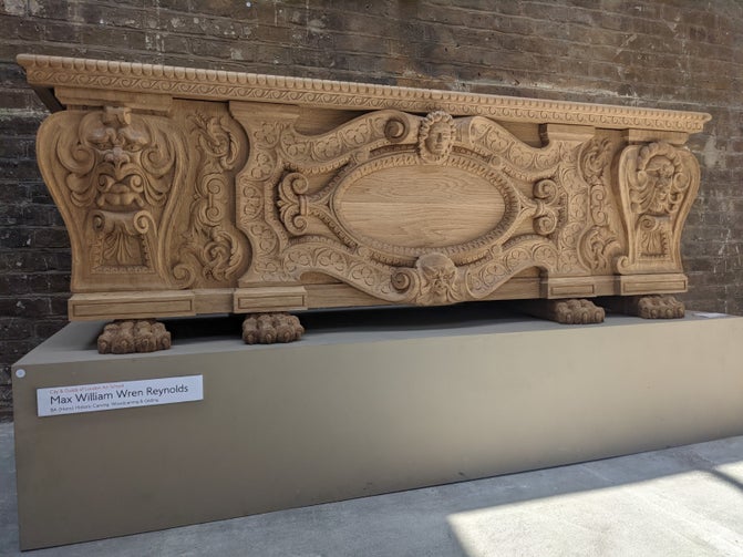 A large wooden chest on a grey plinth. It is elaborately carved, with a pattern and faces.