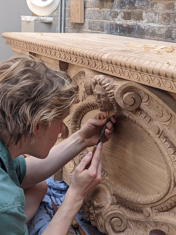 Someone carving a piece of wood on a larger chest. The chest has carved patterns over it.