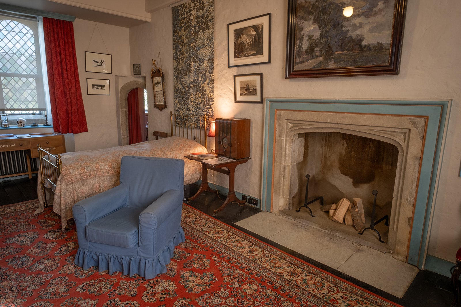 Eddy Sackville-West's bedroom in the Gatehouse Tower, with decorated fireplace, bed with a lightcoloured bedspread on, a blue armchair, patterned red carpet