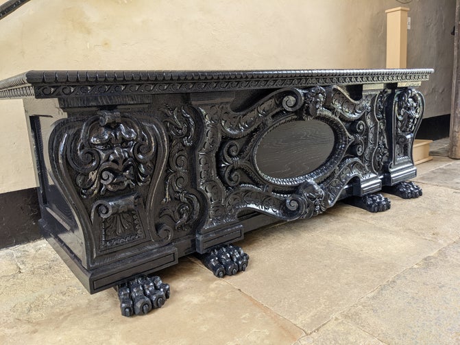 A large wooden chest painted in very dark blue, almost black paint. It is on a stone floor. There are carved patterns and faces on the chest and the feet are in the shape of lion's paws.