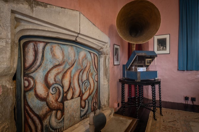Gramophone with a large horn on top of a a blue base, sitting on a black table. It's next to a fireplace that is decorated and painted with light blue and peach patterns.