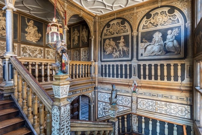 View of a grand staircase with walls painted in grey and white, with wooden stairs and rails, leading to a balustrade overlooking the staircase.