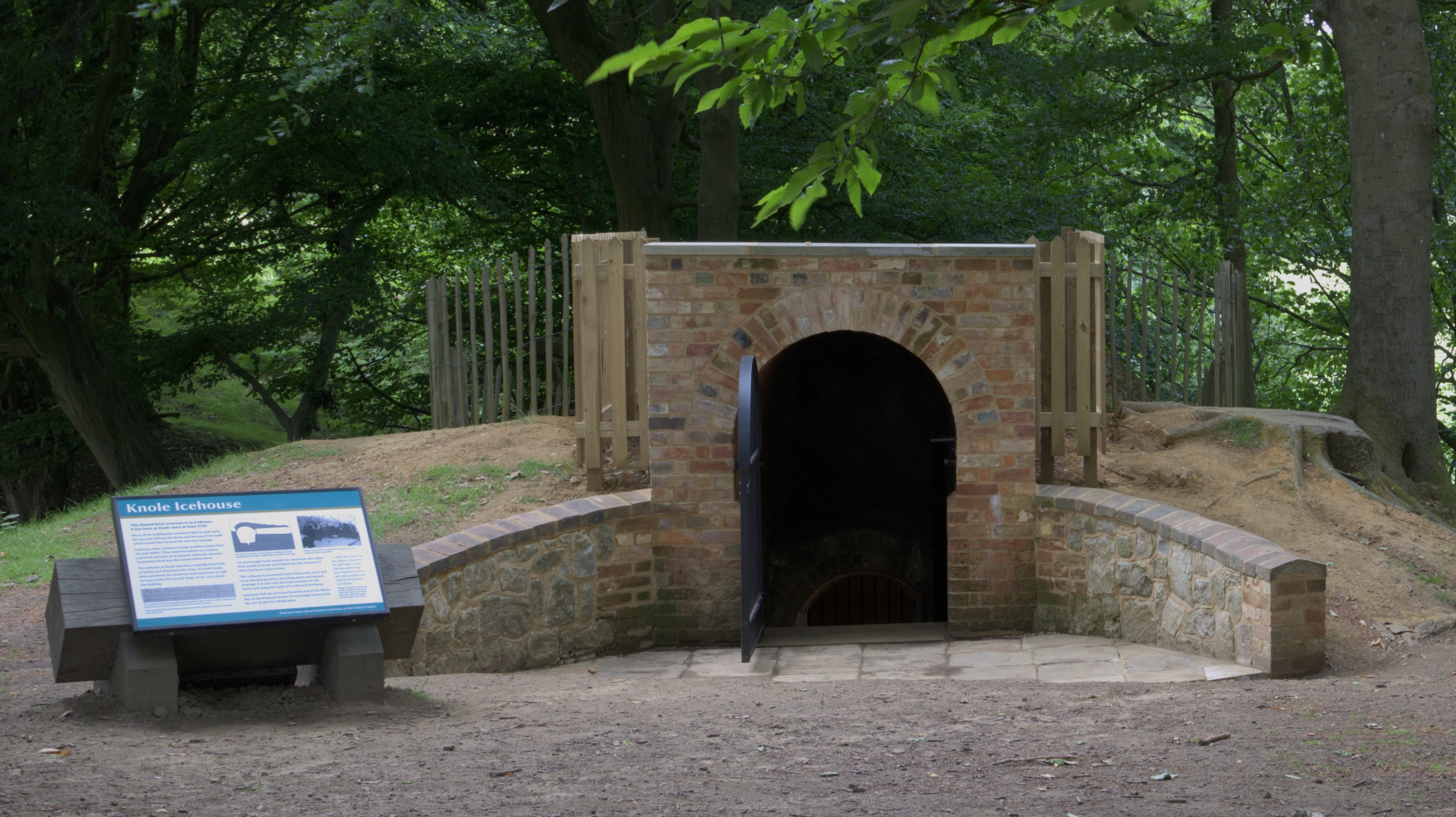A brick fronted structure with an arched entranceway. A black metal gate is open. There is a sign to the left of the structure and trees in the background.