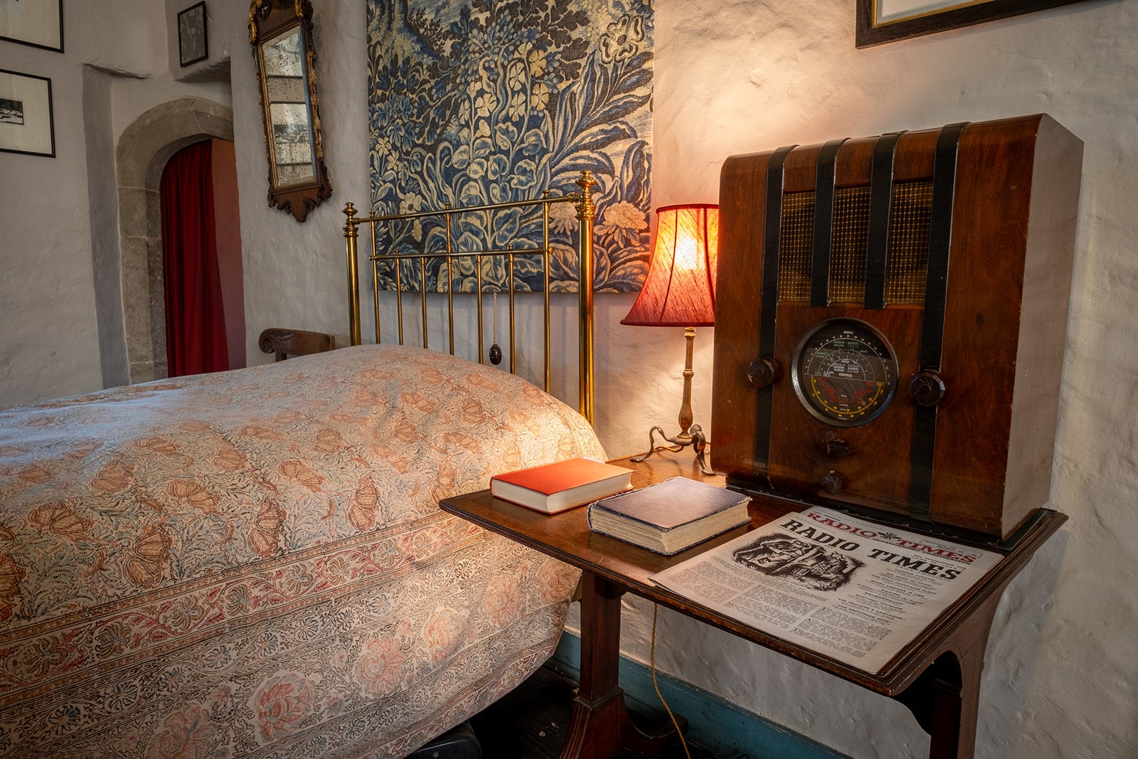 Brown and black radio on  a wooden table next to a bed with a patterned wall hanging behind the head of the bed.