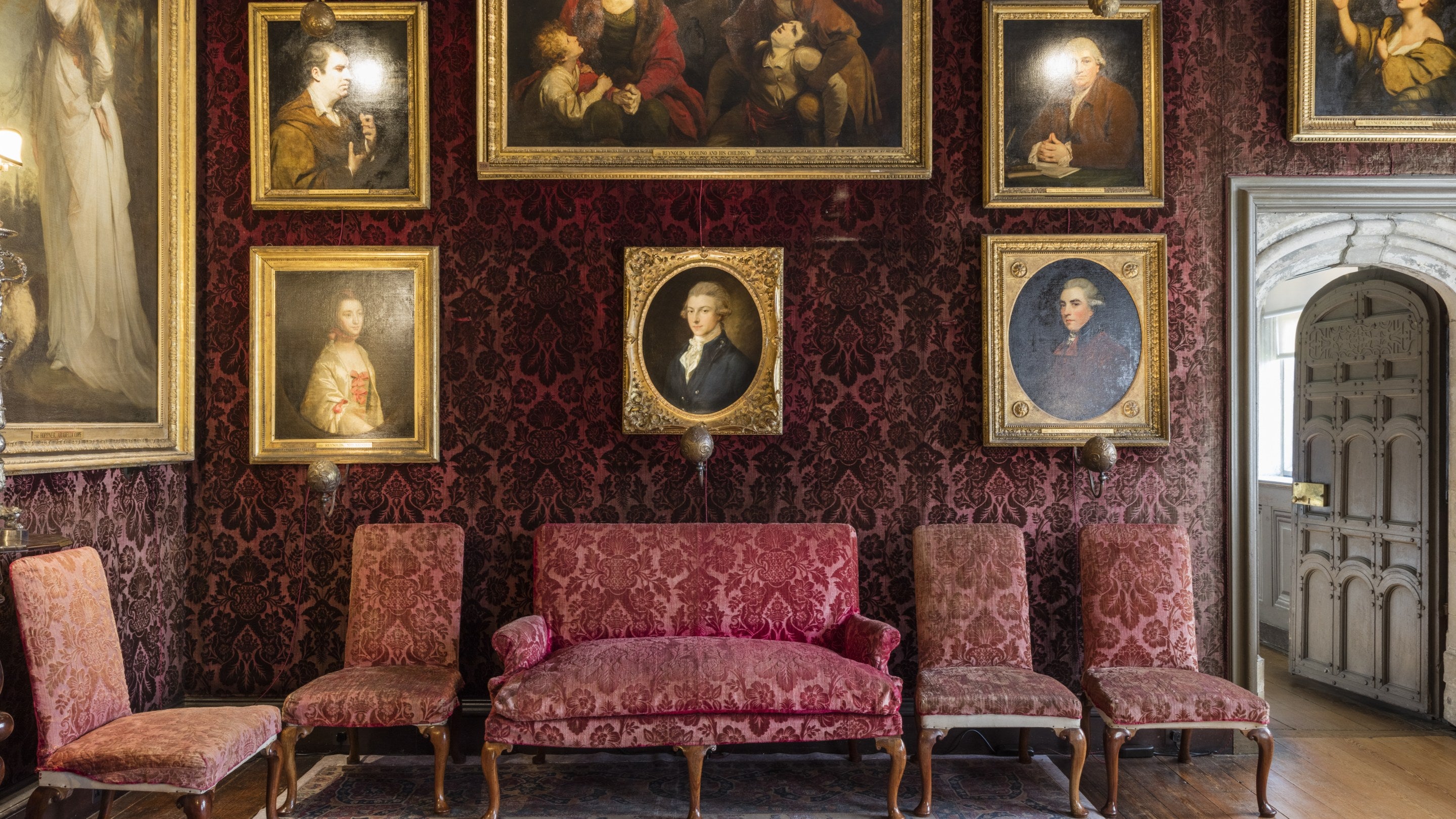 The interior of the Reynolds Room at Knole, Kent, showing a collection of portraits hanging on a richly decorated wall above some chairs.