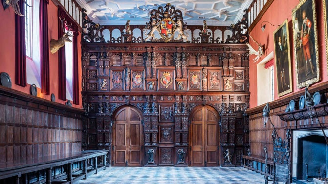 The Great Hall at Knole in Kent looking towards the ornately carved wooden screen across one wall and geometrically patterned white plaster ceiling of squares and octagons