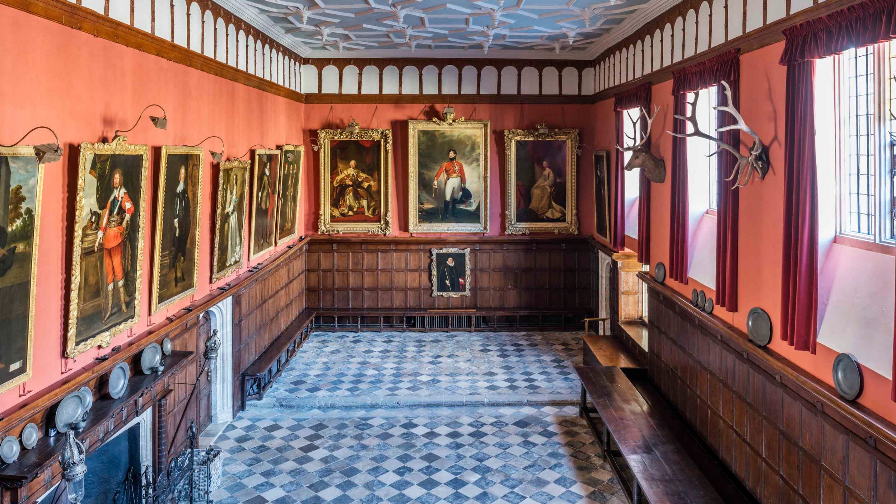 A view of the Great Hall, looking down from the top of the wooden screen gallery at Knole, Kent across a black and white checked floor, dusty red walls and white plaster geometrically patterned ceiling