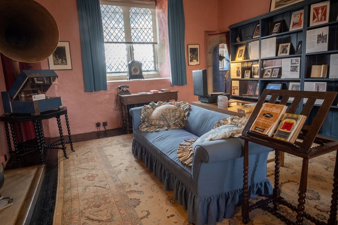 A room with a blue sofa, a large gramophone, a large window and a patterned rug, plus shelves of books to the back.
