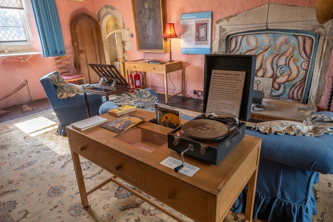 A record player on a light wooden table in a room with a blue sofa, a large window and a patterned rug. There is a large portrait on the wall, a decorated fireplace and the walls are painted in a terracotta colour.