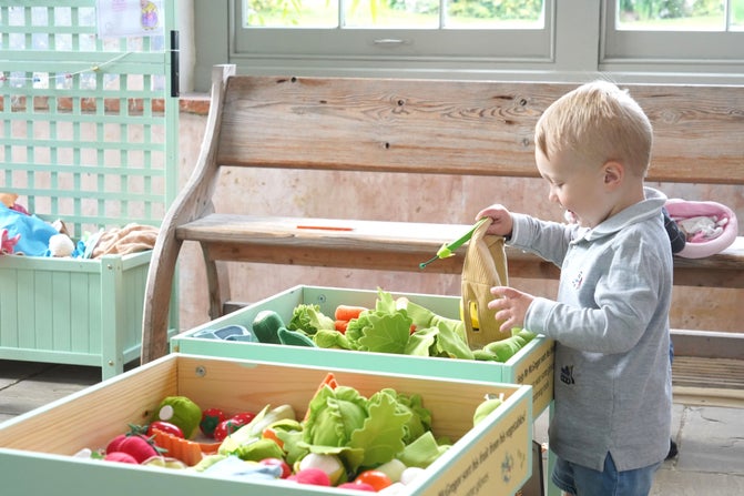 A small child is standing next to a wooden green stand that has vegetable-shaped toys in. They are picking up one of the vegetables and smiling.