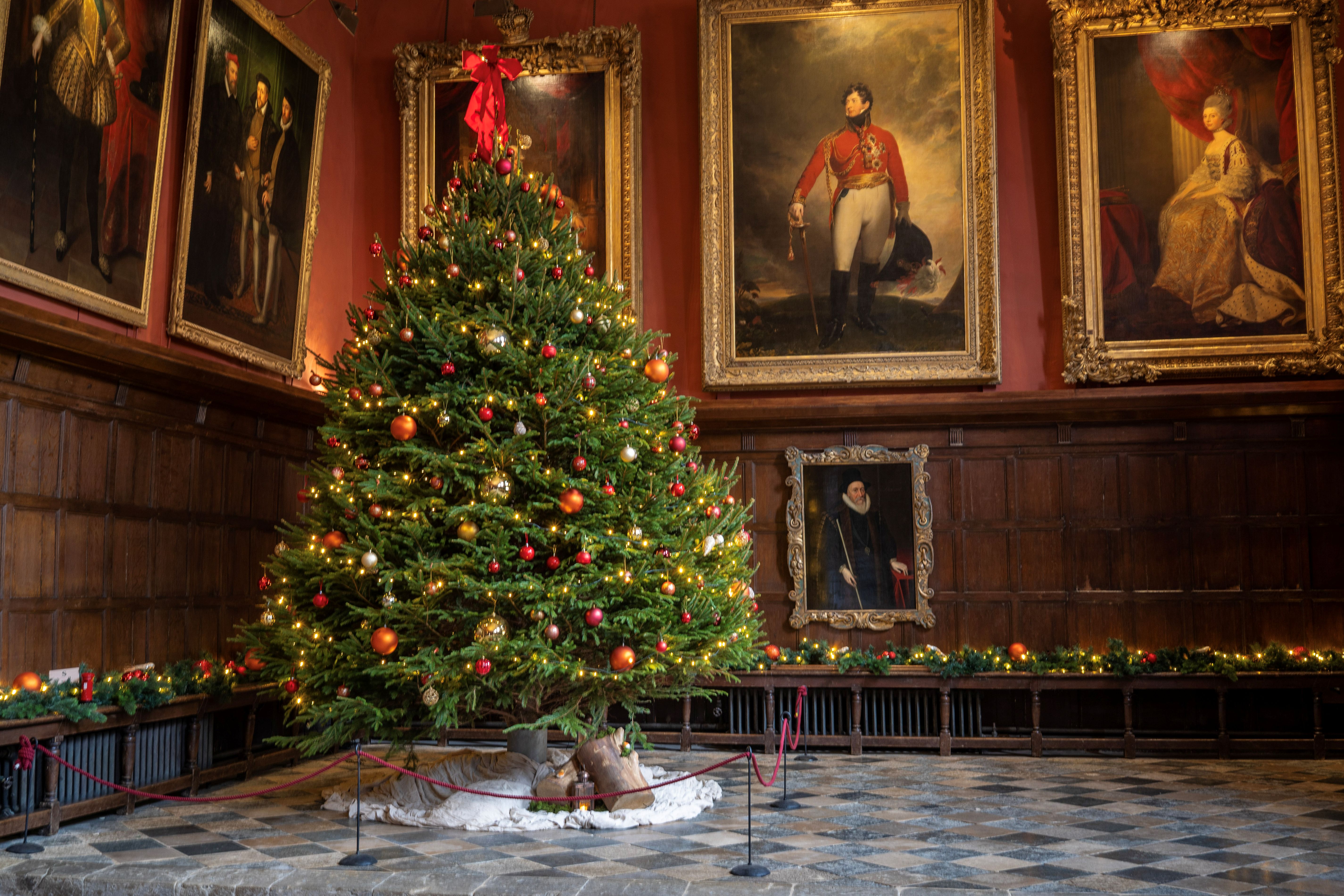 Christmas tree decorated with red, gold and orange baubles, with small lights and topped with a red box. There is a large portrait paintings on the walls either side of the tree. There is wooden panelling below these paintings, a wooden bench with a green foliage garland laid along it, and a chequered stone floor.