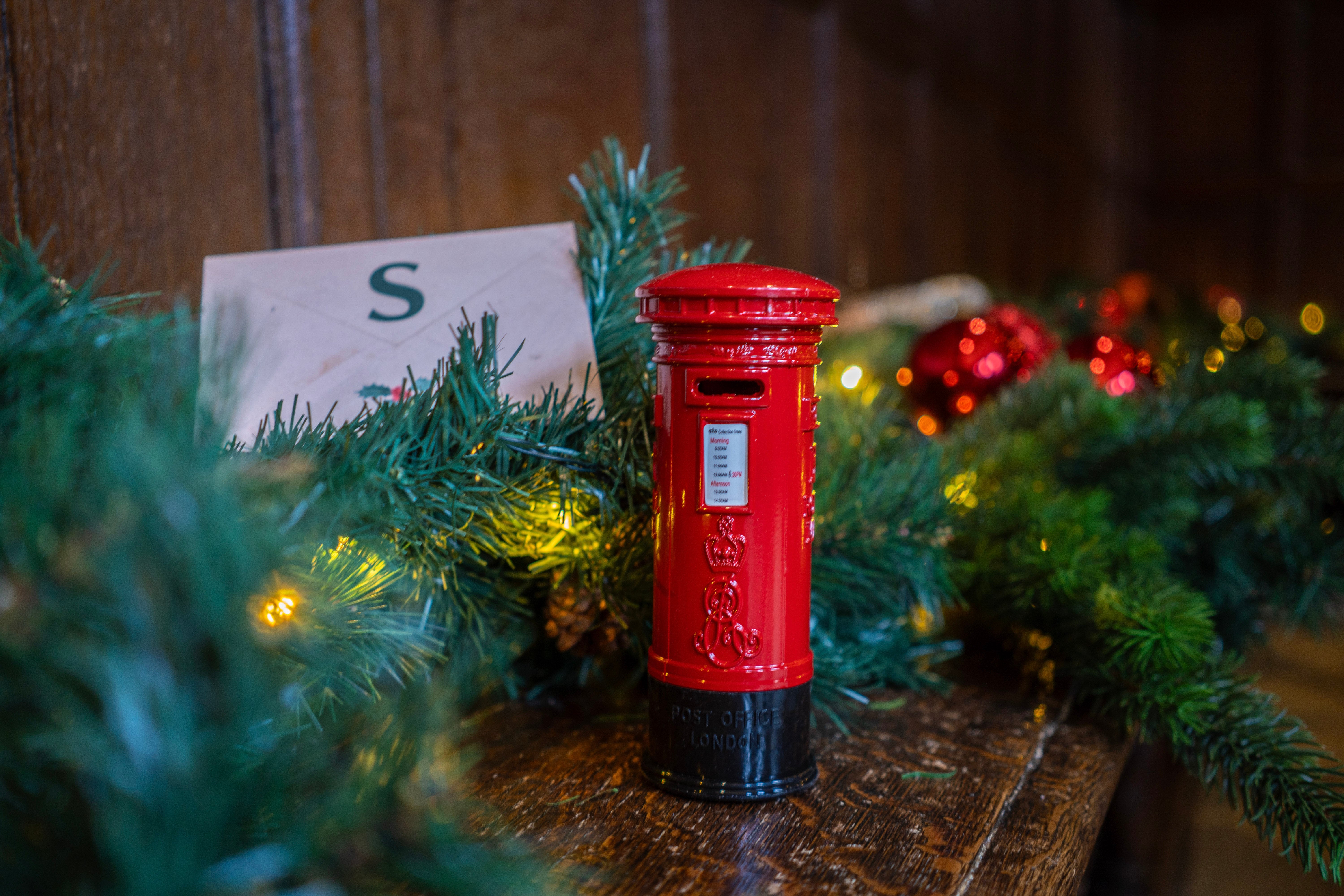 A small red postbox on a wooden bench with green foliage around it, and an envelope next to it with the letter S on.