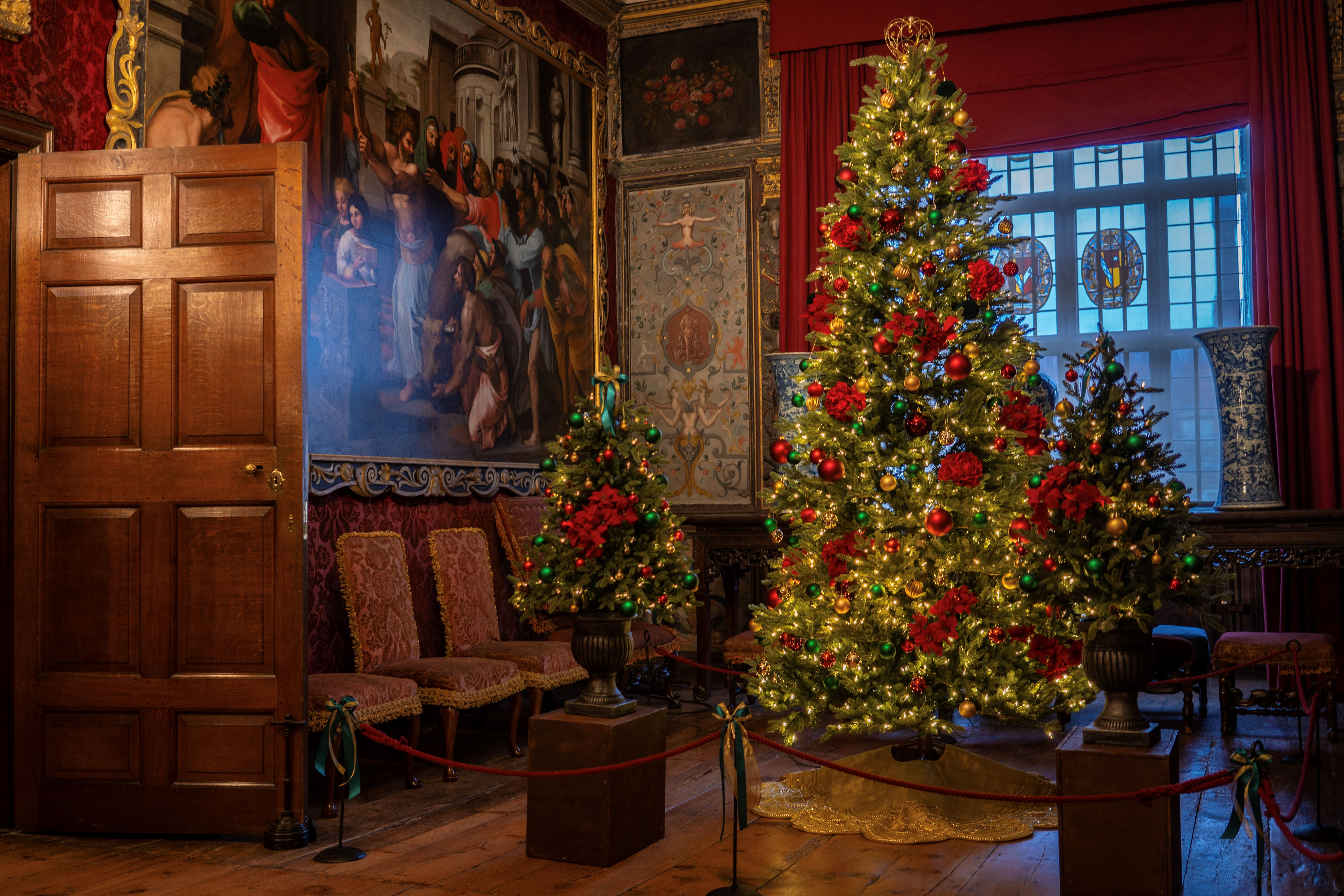 Christmas trees decorated with baubles, red flowers and bows and small lights. There is a large painting on the wall on the left behind an open wooden door, and a large window in the background.