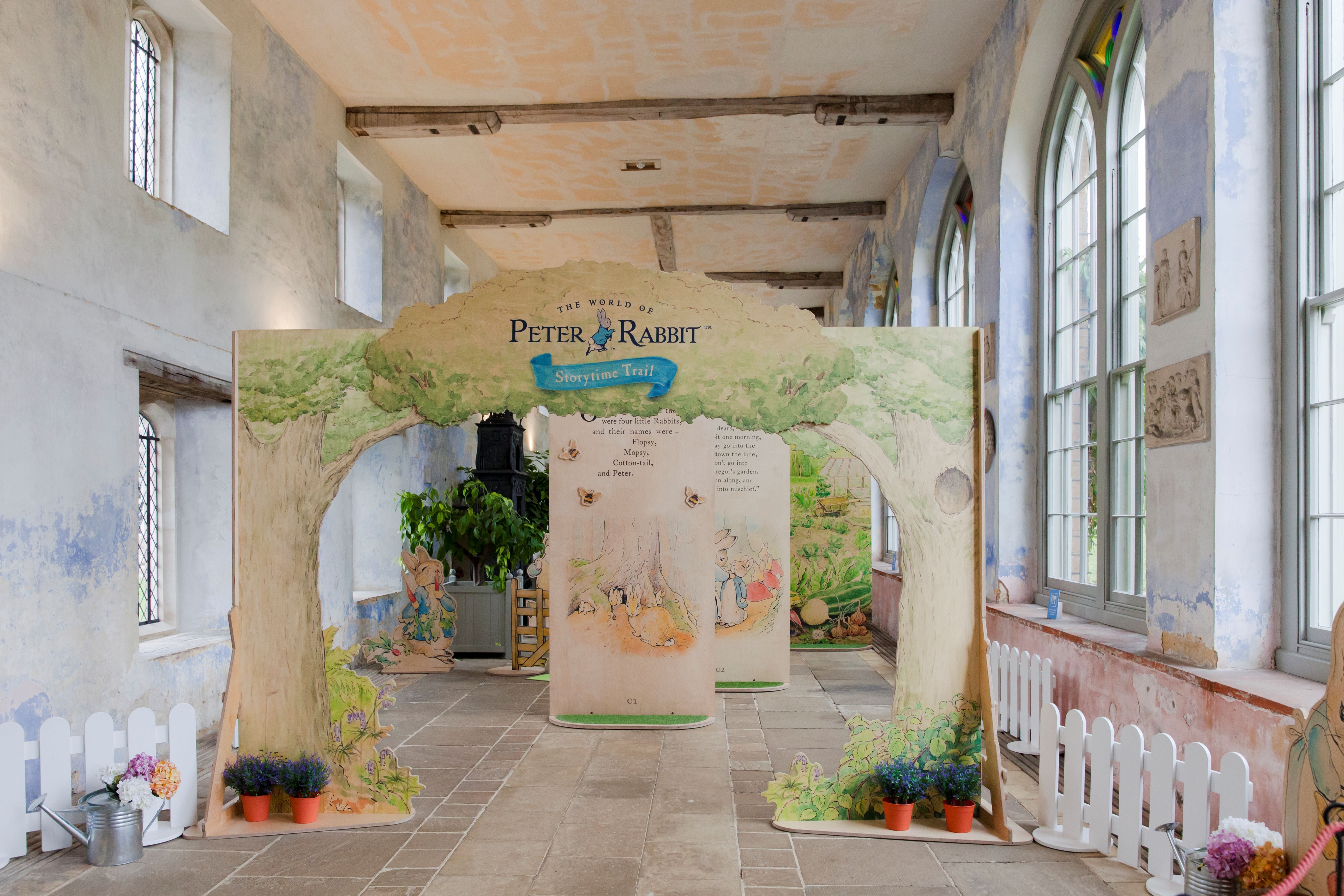A green illustrated wooden archway in a room that has light grey painted walls and tall windows. There are more illustrated wooden boards behind the arch, white fences, pots with flowers in and a tiled floor.