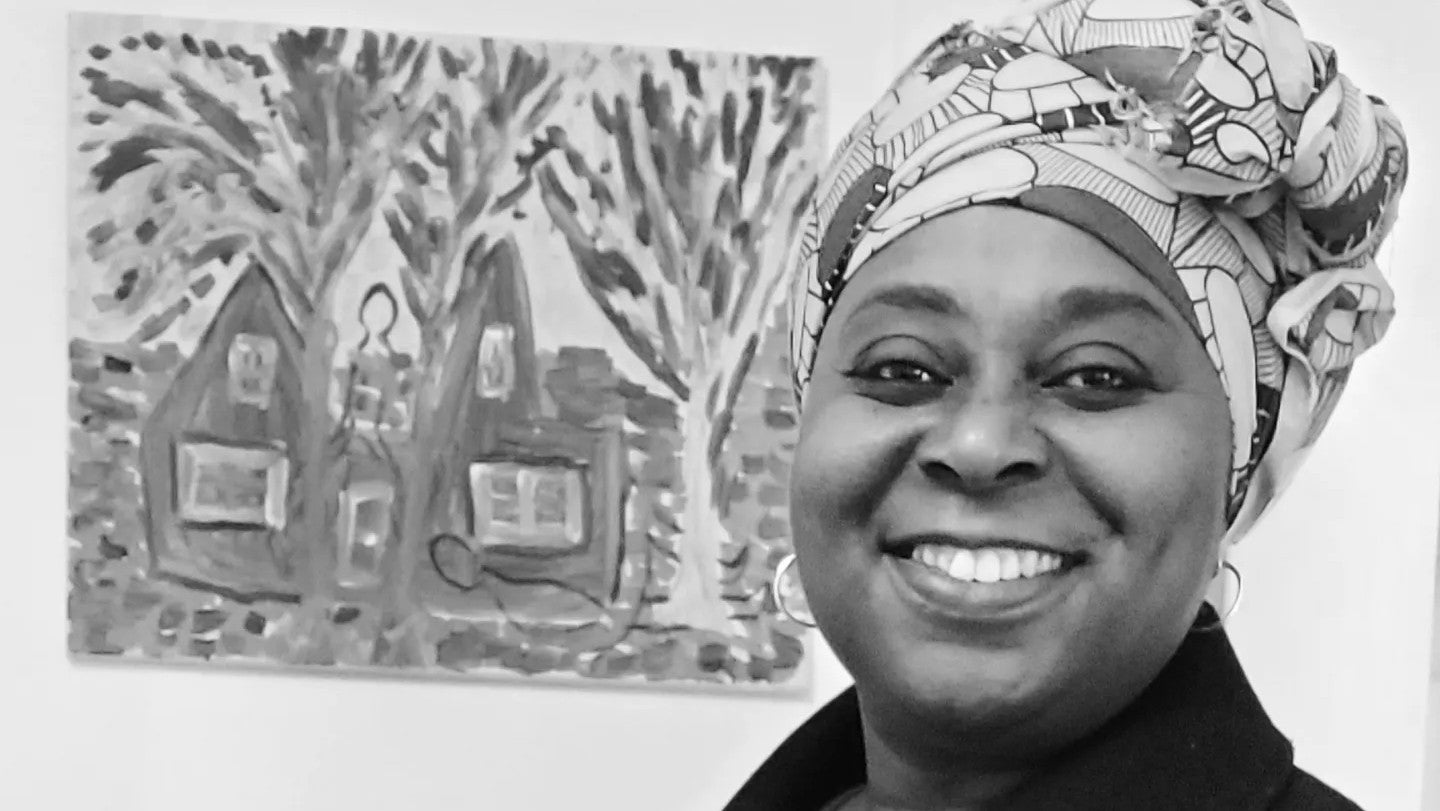A close up photo, in black and white, of a smiling person standing next to a painting of a house.