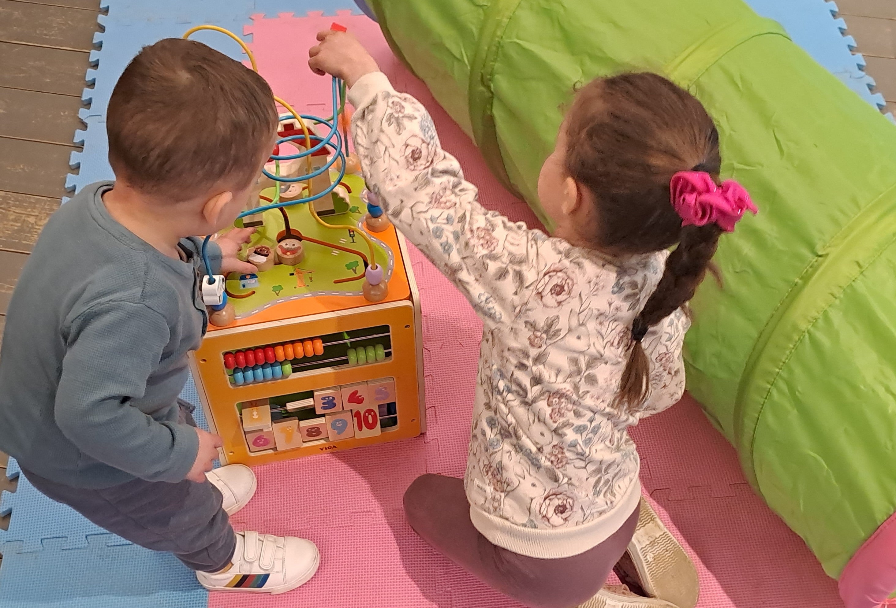 Two young children playing with a wooden toy on pink and blue mats with a colourful play tunnel next to them.