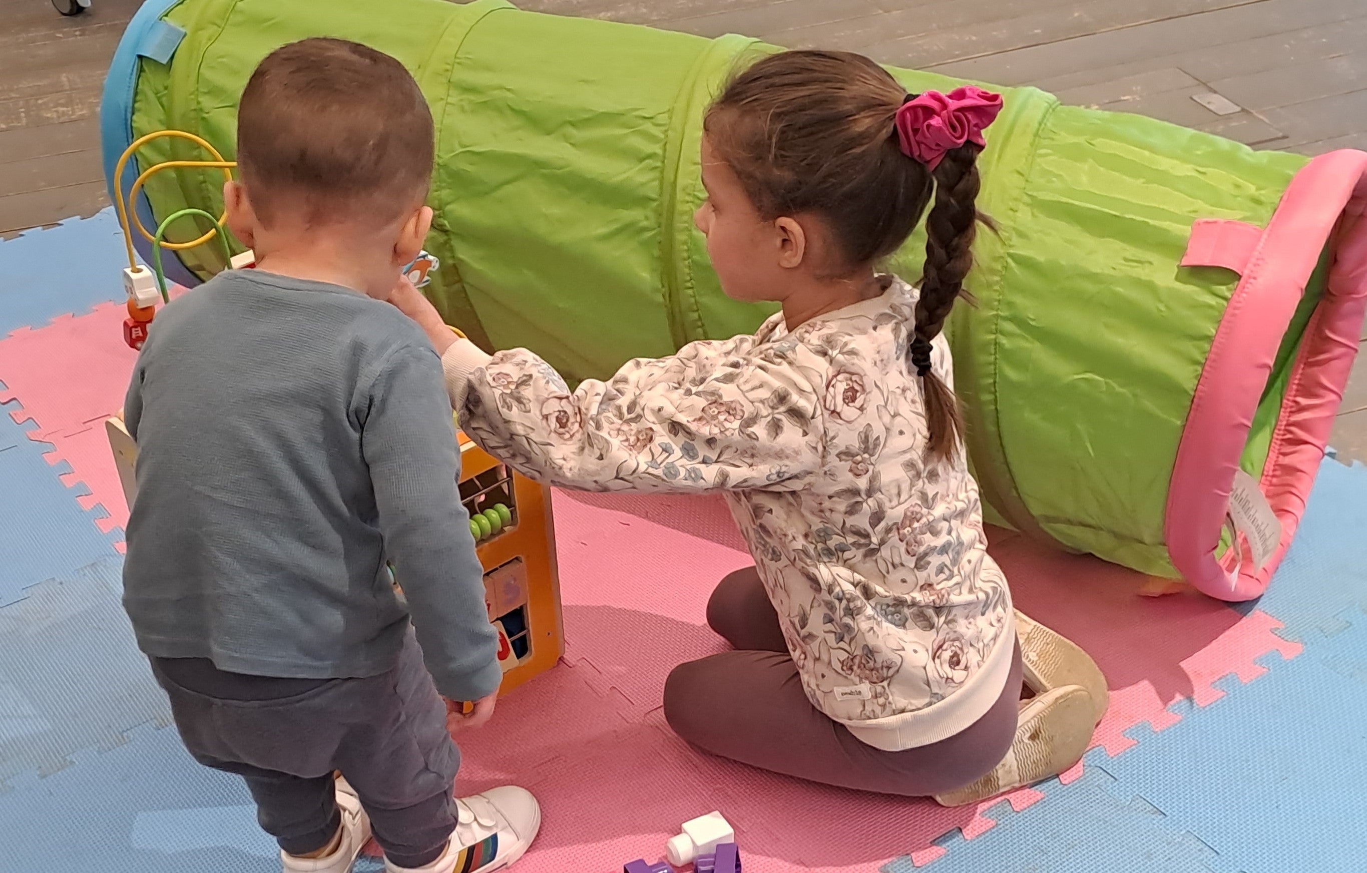 Two young children playing with a wooden toy on pink and blue mats with a colourful play tunnel next to them.