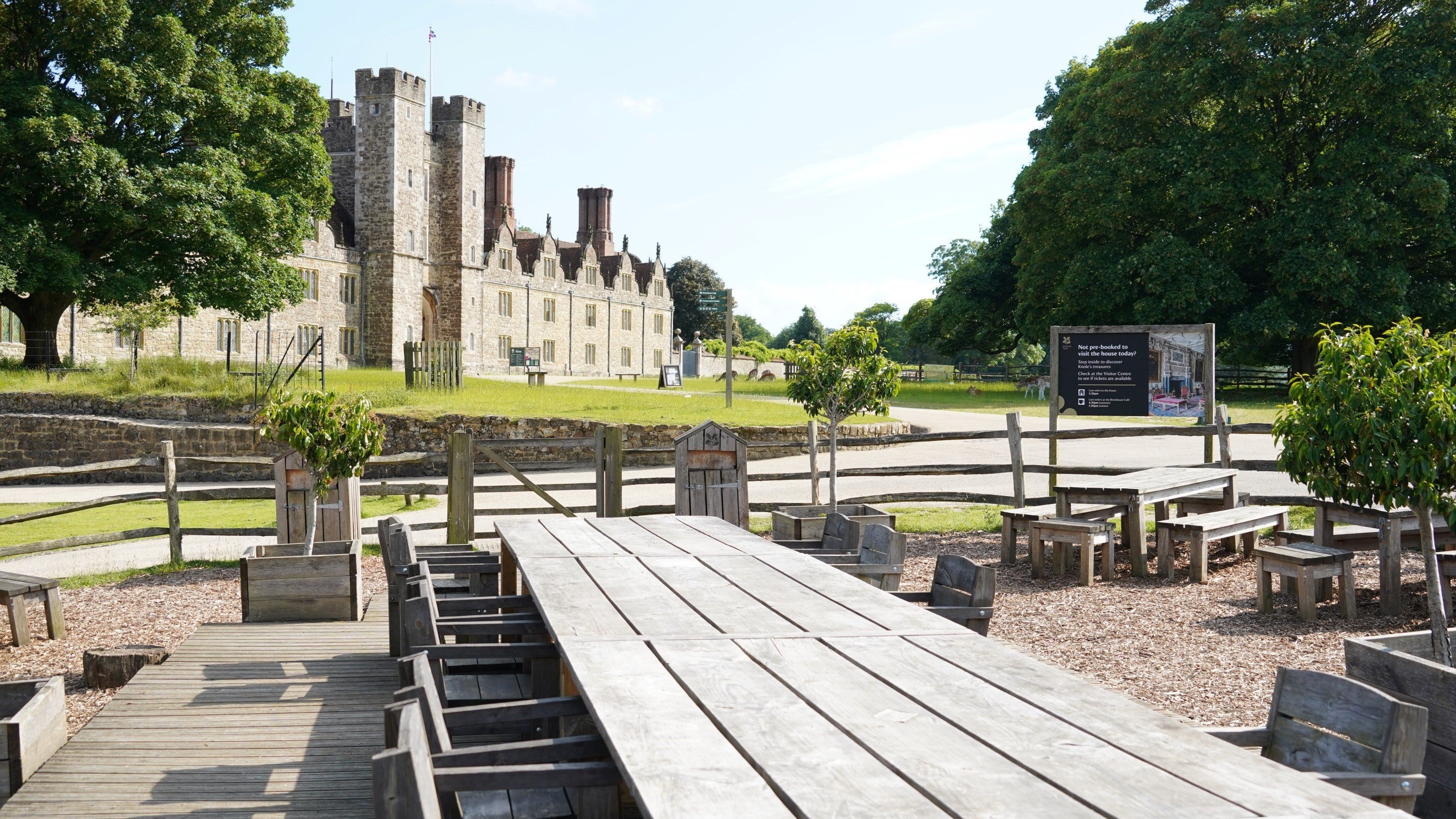 Picnic area with wooden tables, benches and chairs, with historic building, grass and green trees in the background.