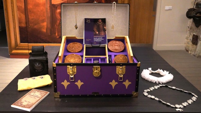 A large purple box on a grey table, with the lid open to reveal contents, which include round, dark red discs, a white circle of material, a necklace and a black lamp.