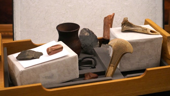 A selection of items aid in a wooden box, including tools made from wood and stone, a metal buckle, and a cup.
