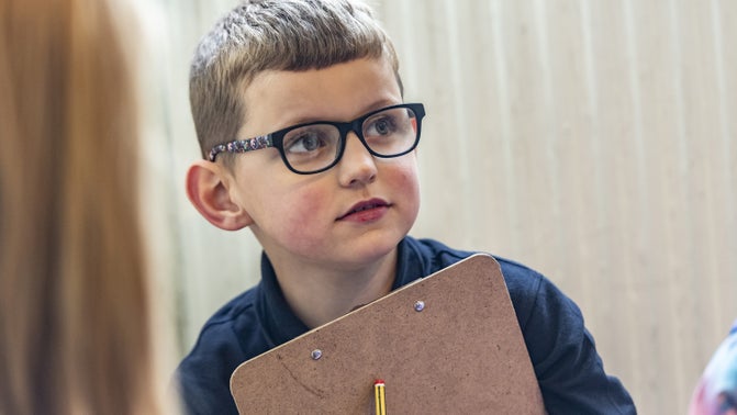 Child looking into the distance and holding a brown clipboard in front of him, with a pencil in one hand.