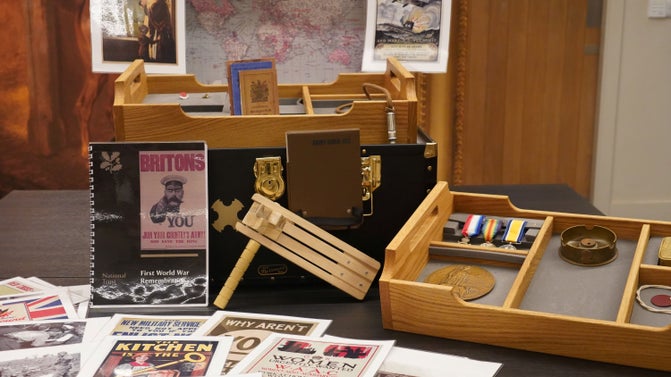 A large box on a grey table with various items in it and laid around it, including posters and printed material, a brown covered notebook, replica medals, and wooden ratchet rattle.