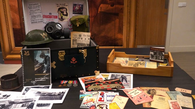 A large box on a grey table with various items in it and laid around it, including posters and printed material, replica gas mark and helmet.
