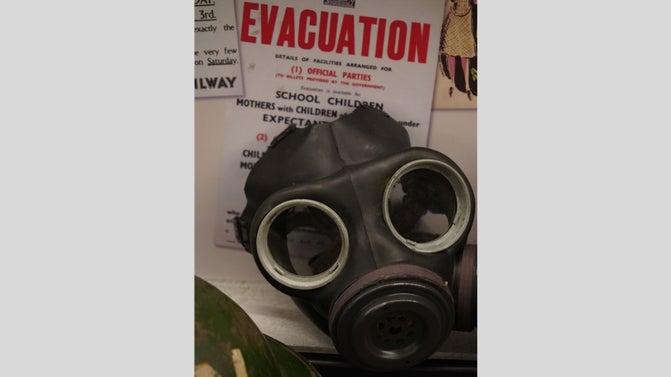 Black gas mask with large eye features with a poster behind it that says evacuation in large red text