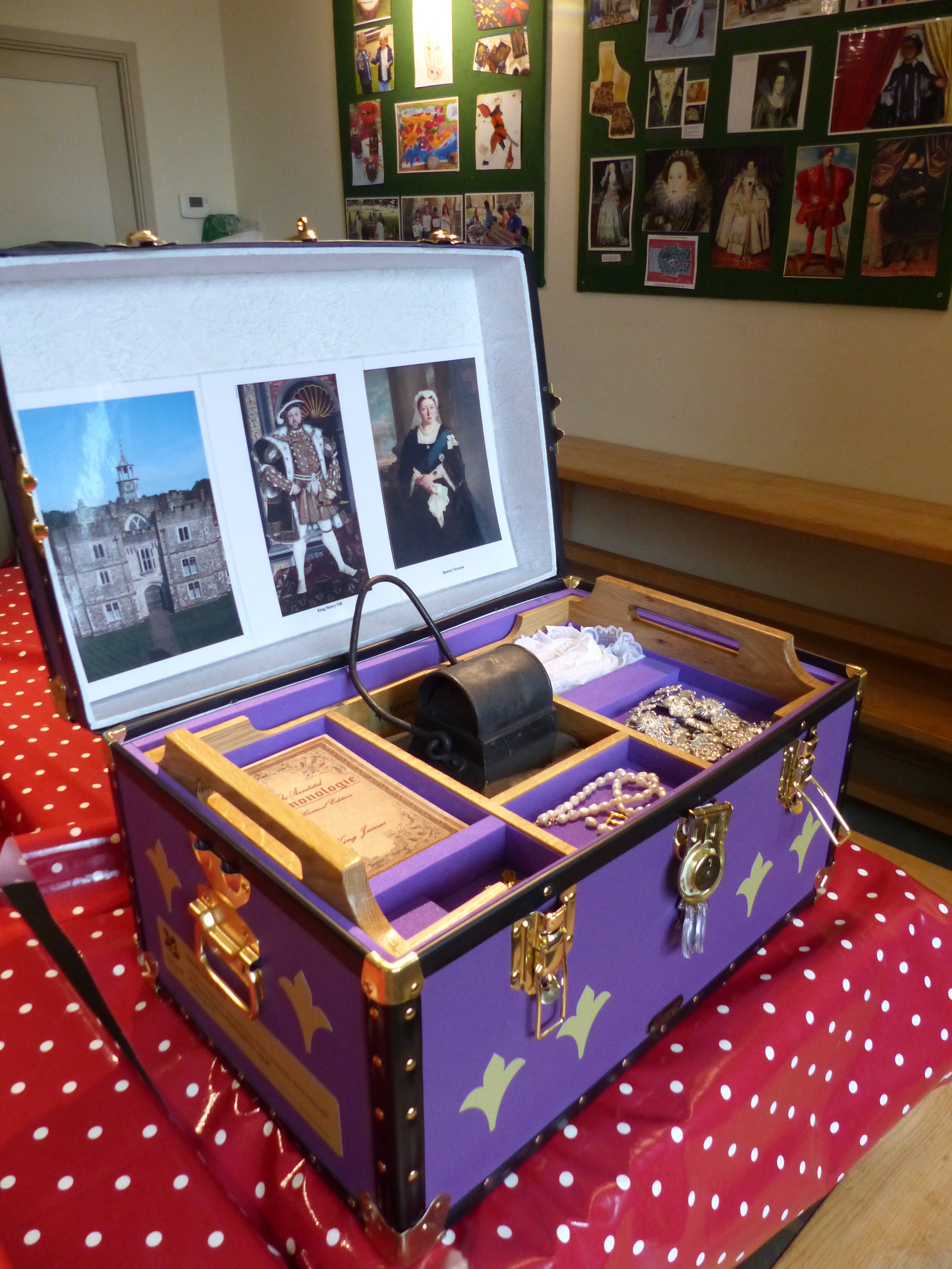 Large purple case on a table that has a red and white tablecloth on. The lid of the case is open and there are photos and items inside, including jewellery. There are photos on the wall in the background.