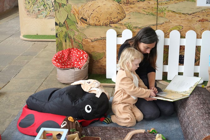 An adult and child are sitting on the floor looking at an open book. There is a large soft toy ladybird next to them and a white fence placed behind them.