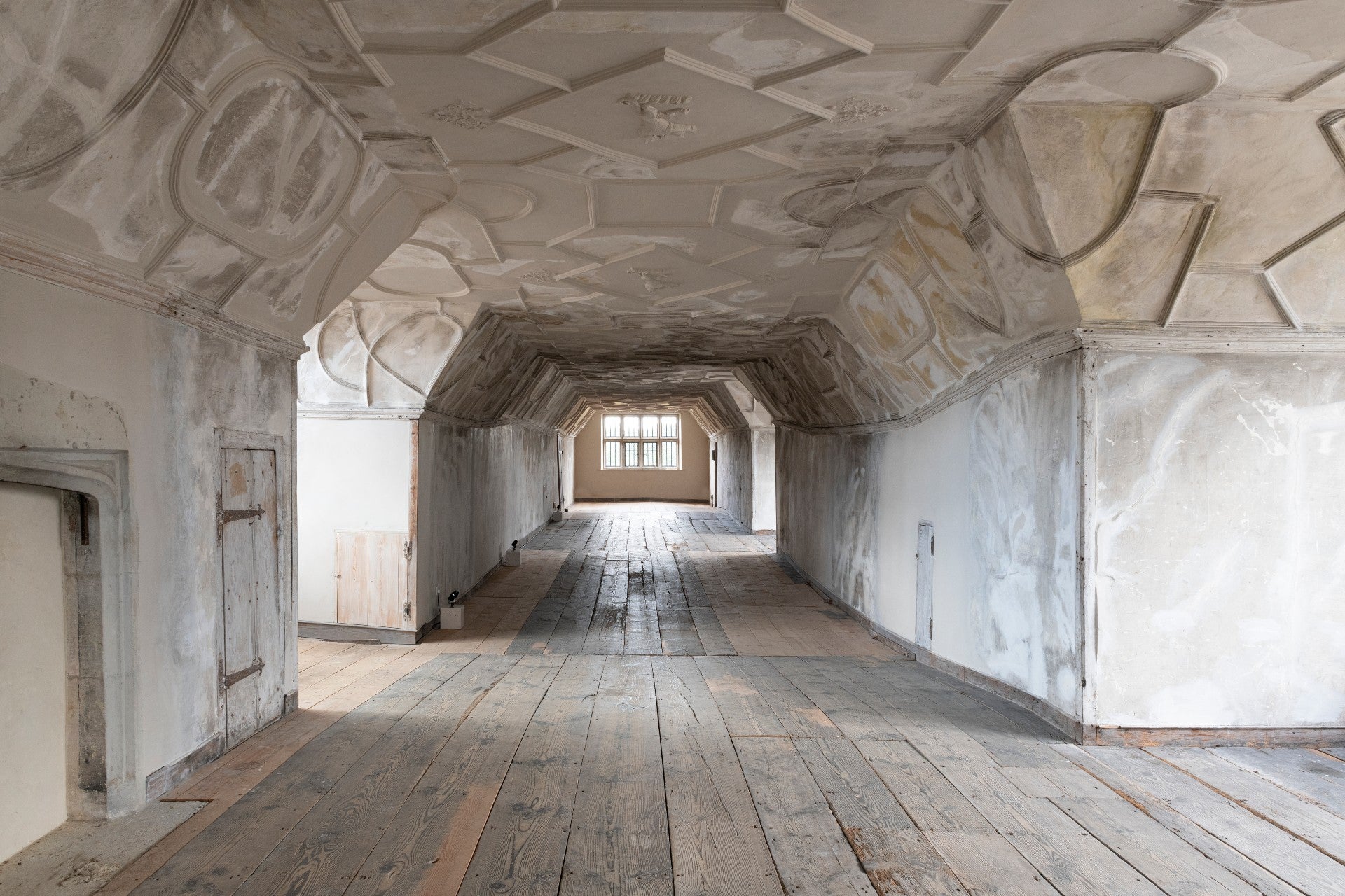 Long view down a covered area with a white ceiling, wooden floorboards and a window at the end.