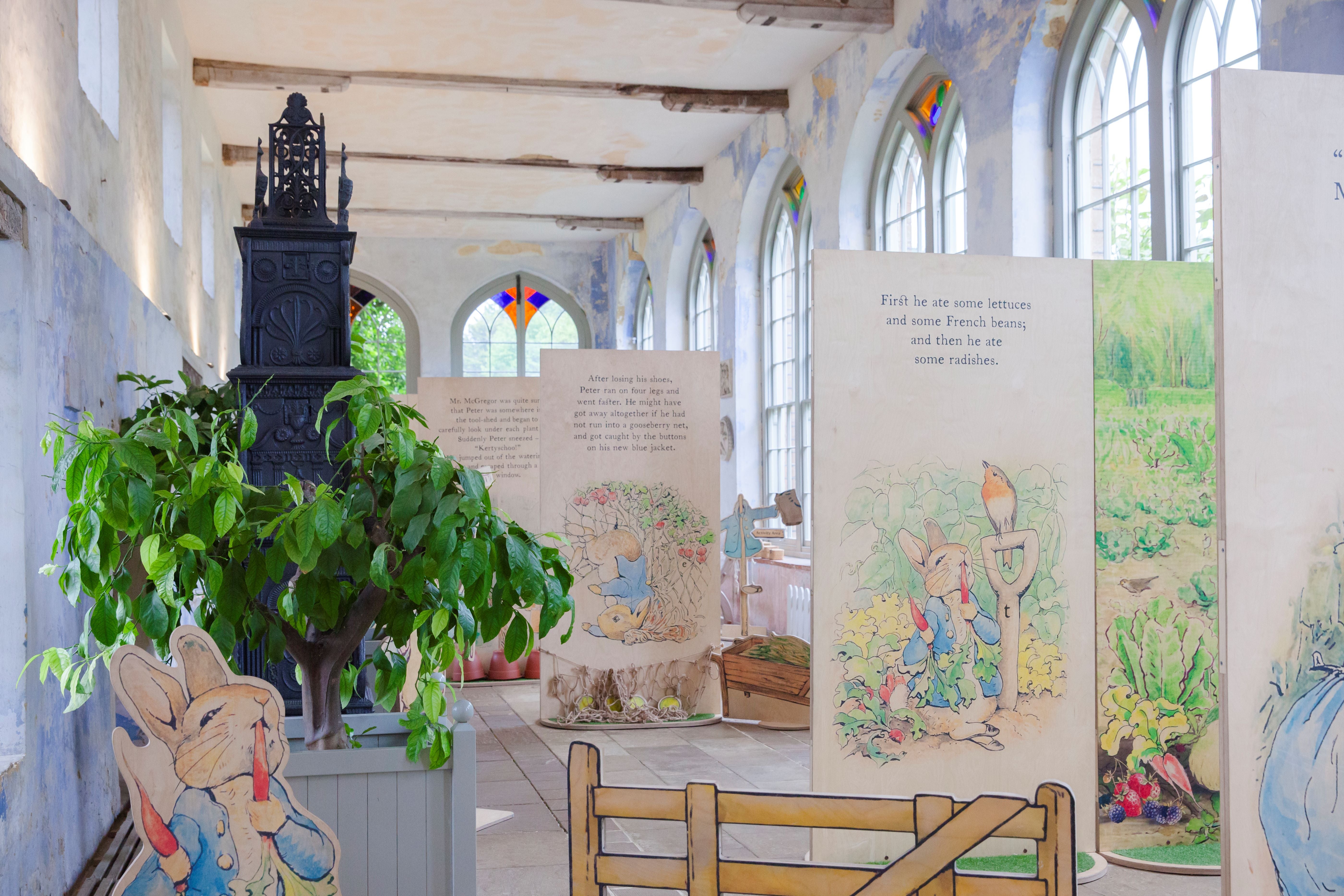 Illustrated wooden boards in a room that has light grey painted walls and tall windows. There is a tall black metal object to the left, a green leafed tree, and a wooden cut out of a rabbit eating a radish.