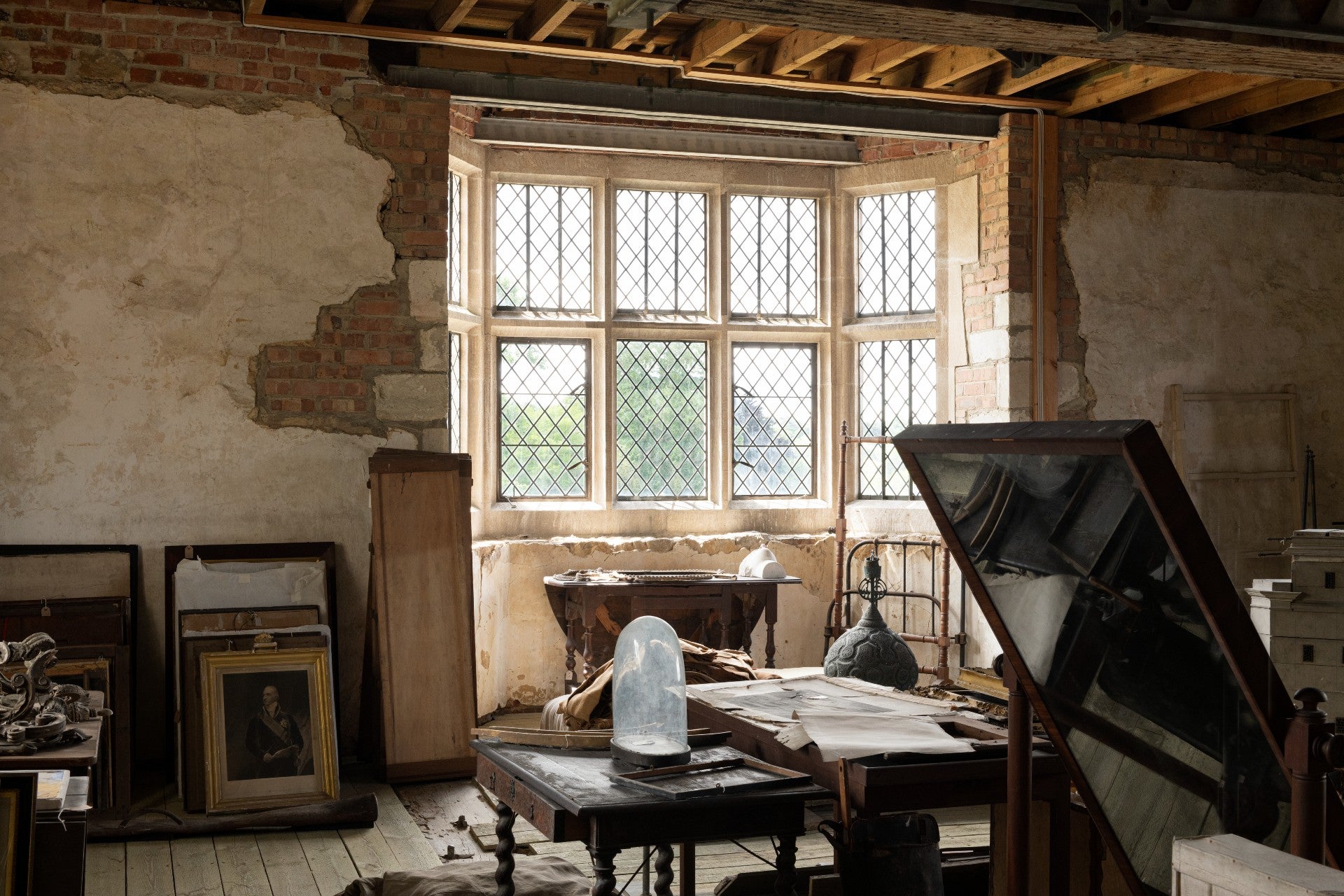 View into a room with furniture including chairs, paintings and a mirror in it. Some of the walls are showing exposed brickwork. There is a large bay window in the middle of the scene and a beamed ceiling.