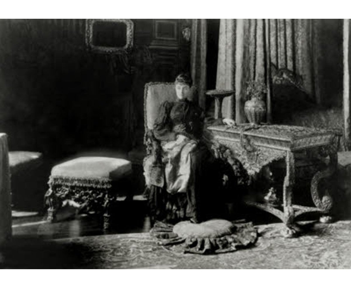 A black and white image of a woman seated next to an ornate table and a cushioned stool. There is a curtained bed in the background and a cushion laying on the patterned carpet on the floor.