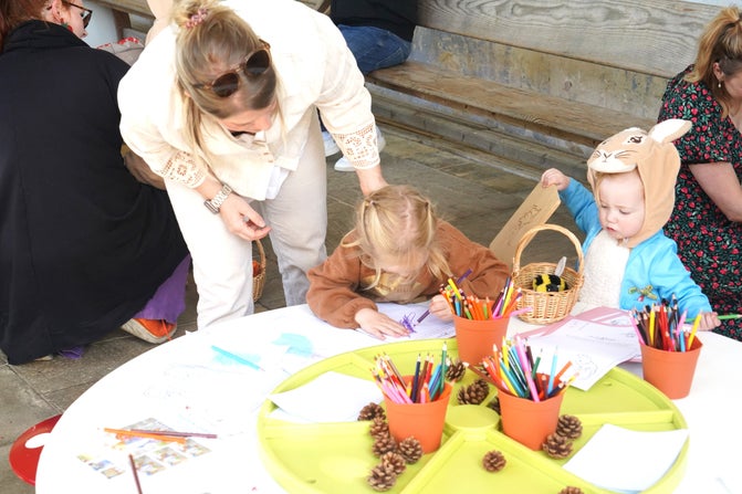 An adult is leaning over two children who are sat at a table with colouring pencils and activity sheets on. The children are dressed up in rabbit costumes.