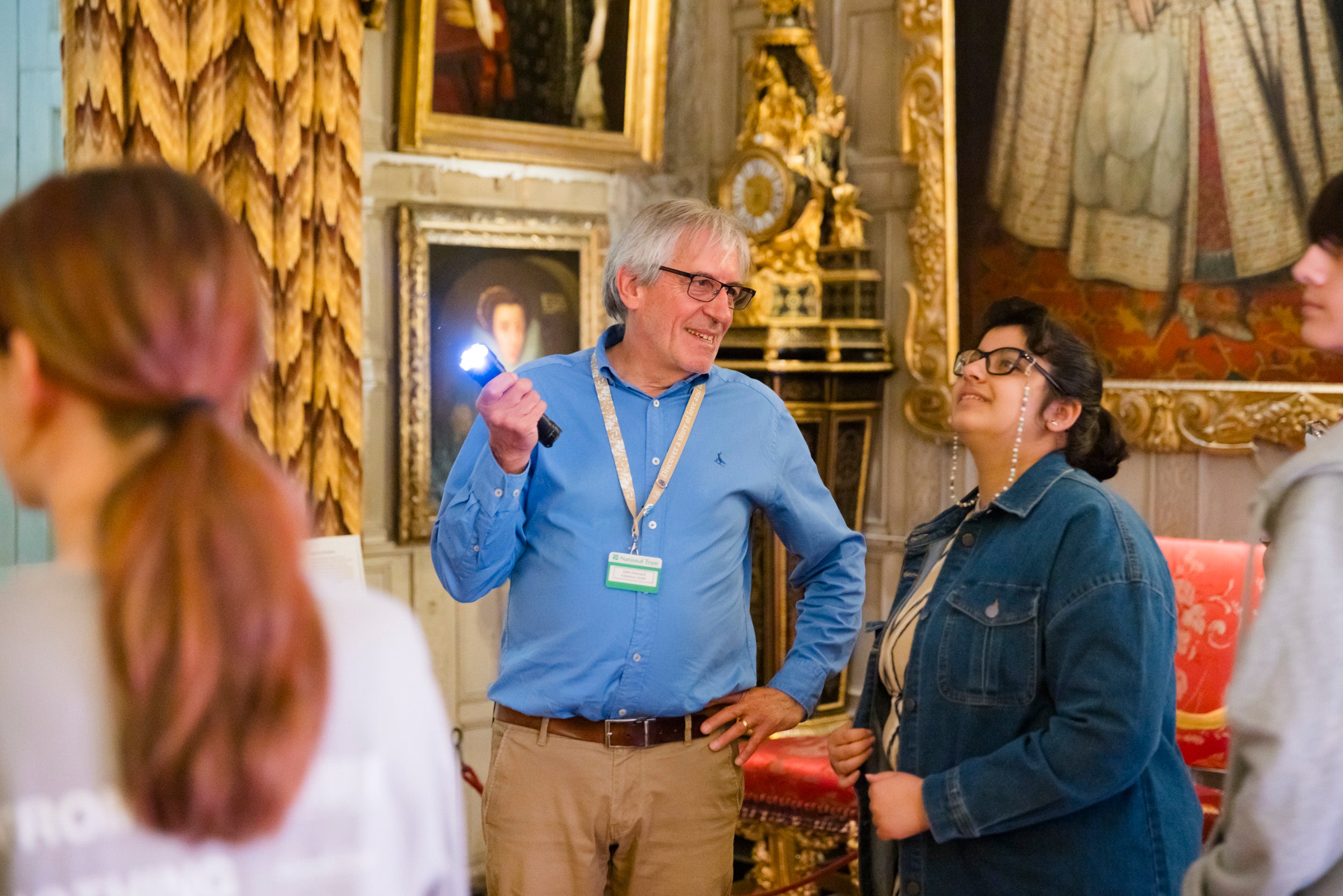 Visitors standing in a room that has paintings on the wall and gold coloured curtains. One person is holiding a torch, to show something to the other person. There is a ornate gold clock in the background.
