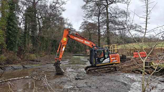 Image of an excavator working to remove silt from a drained pond bed, surrounded by trees on a cloudy day