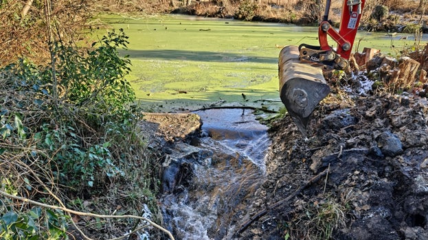 Image of water draining from a weed-covered pond after an excavator removed the old outflow system on a sunny day with the excavator bucket alongside the outflow