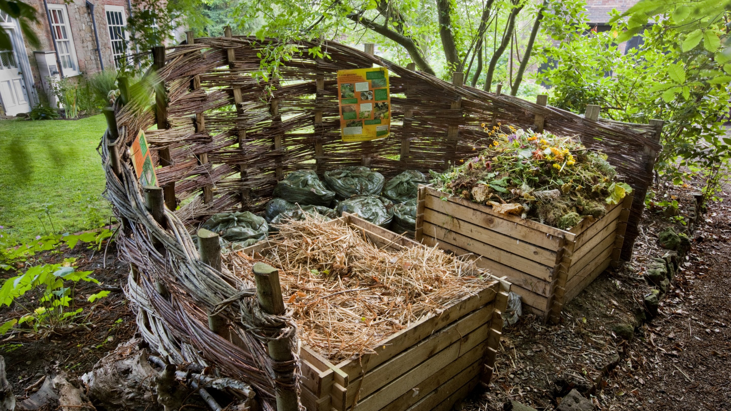 Two wooden compost bins filled with straw and plant waste, surrounded by a wicker fence