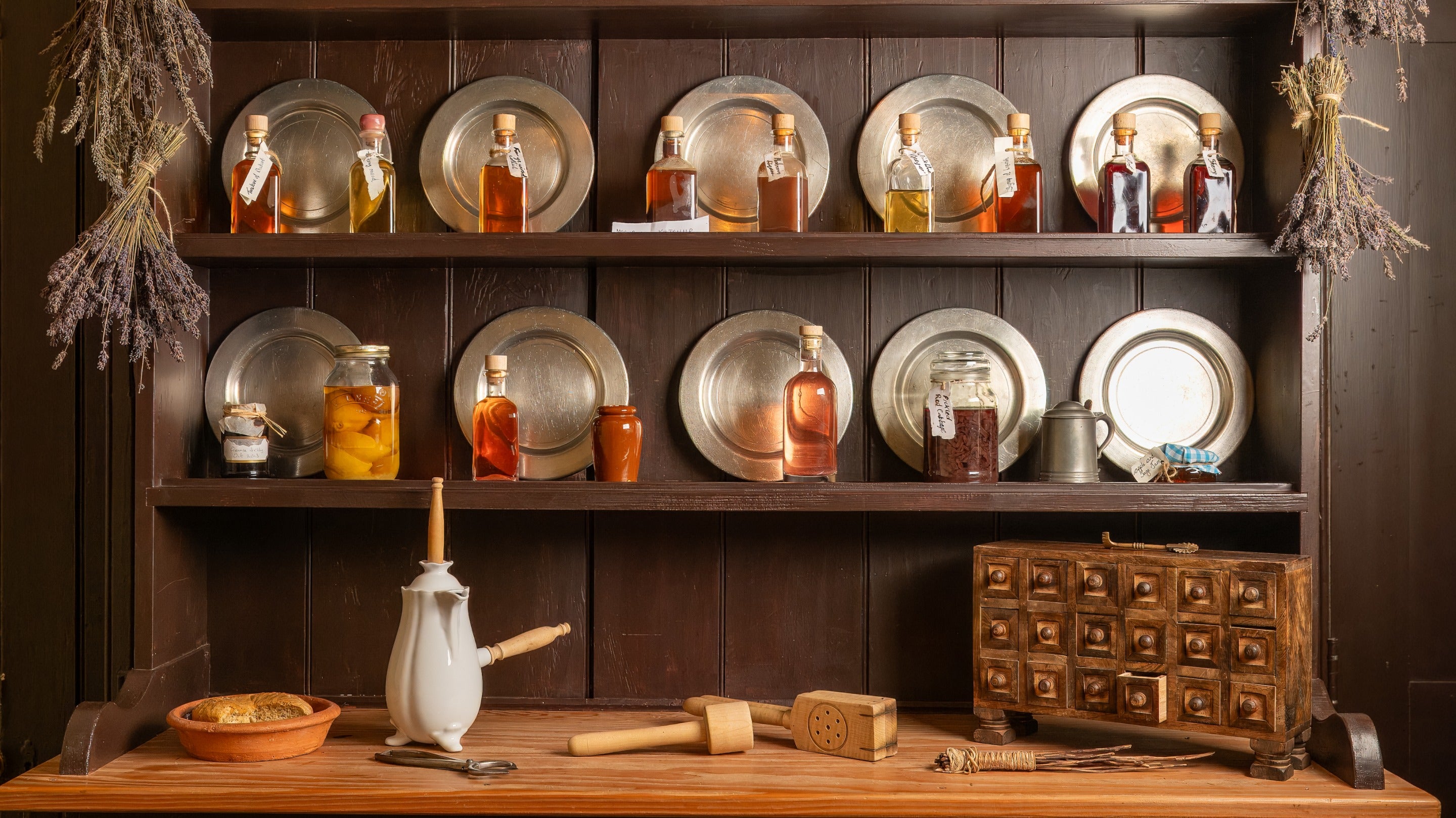 Wooden pantry shelves at Quebec house lined with pewter plates, bottles and containers of preserved ingredients, spice drawer and cooking implements on the work surface and lavender bunches drying on the sides