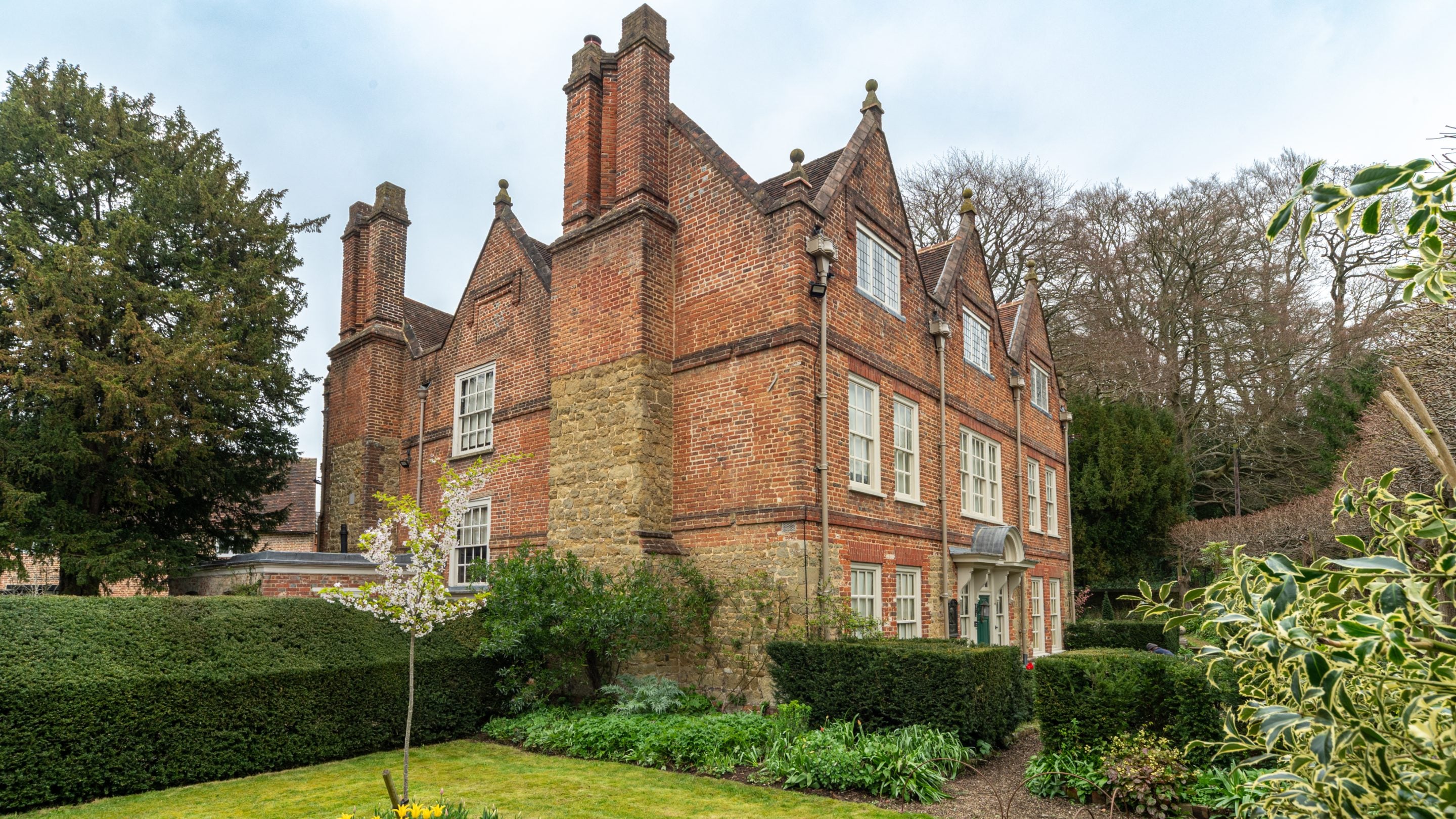 Quebec House in spring. A red brick and grey stone house with several pointed roofs topped with decorative stone spheres, two double chimney stacks and windows on all 3 floors. A young cherry tree blossoms in the garden, surrounded by tightly clipped hedges and other green foliage. A path through the hedges leads to a green front door with a decorative surrounding.