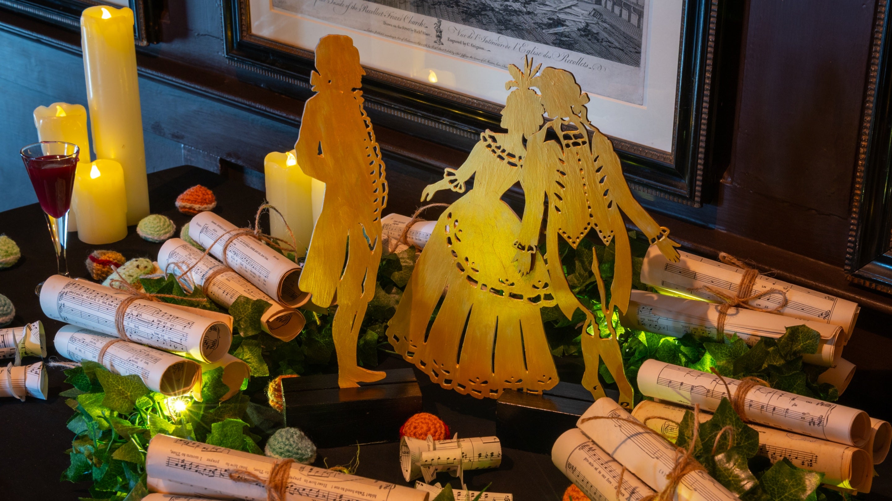 Silhouettes of Georgian era figures amongst a Christmas display of ivy, candles, a glass of port, scrolls of classical music, and crocheted macarons.